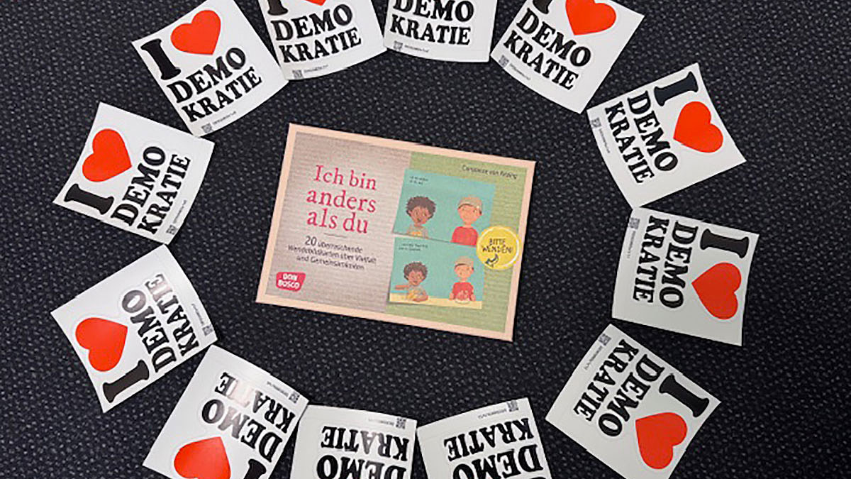 Eine Broschüre mit dem Titel Ich bin anders als du liegt umringt von Aufklebern mit der Aufschrift I love Demokratie. 