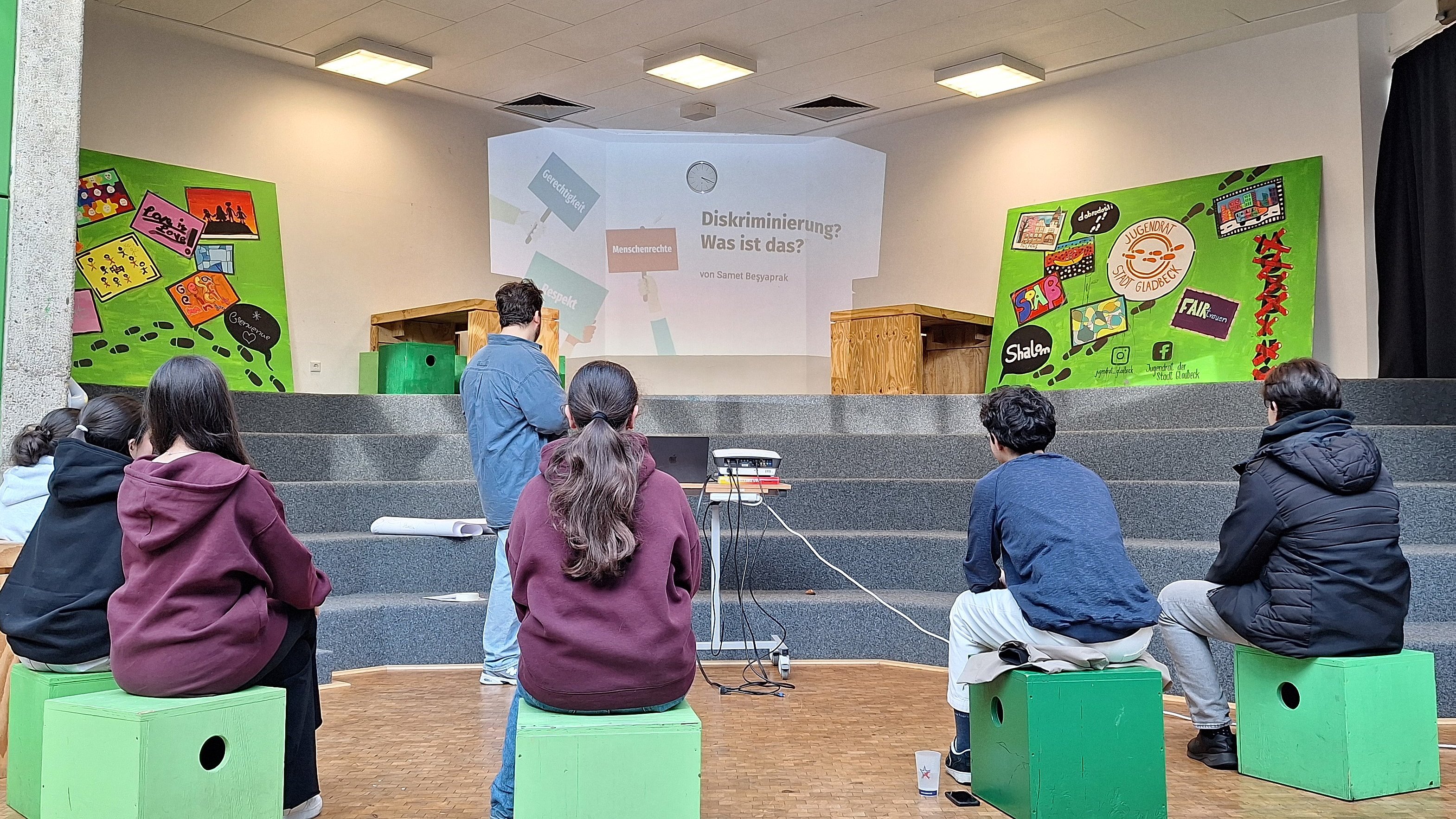 Kinder sitzen auf grünen Klötzen und schauen auf eine Leinwand, auf der ihnen eine erwachsene Person eine Präsentation zeigt.